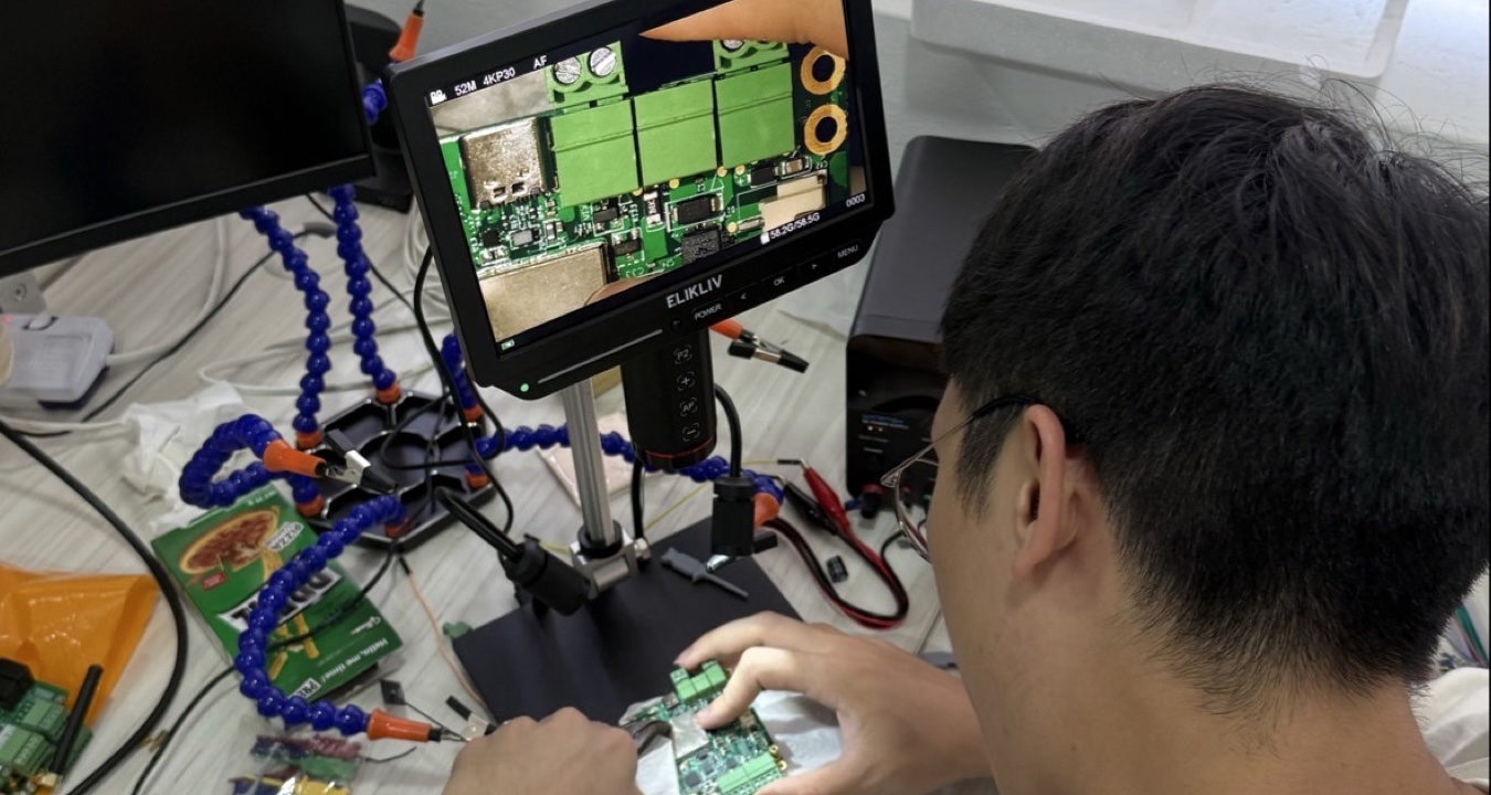 Vantage Point hardware consultant performing close inspection of an IoT PCB under a digital microscope at the testing bench, with helping-hands clamps, probes, and additional development boards in view.
